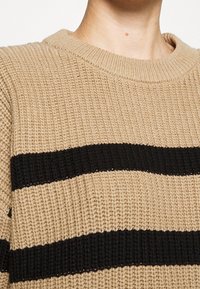 Beige gebreide trui met zwarte horizontale strepen, voorzien van een geribbelde ronde hals en een kenmerkende textuurweving.