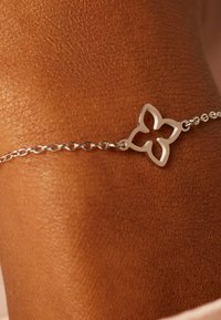 Bracelet en chaîne argentée avec un délicat motif de fleur découpé, reposant sur une peau brune texturée. Les maillons de la chaîne sont fins et bien espacés.