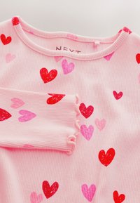 Maglia rosa a coste con maniche lunghe, con motivi di cuori rossi e rosa e polsini a bordo ondulato, piegata per mostrare il dettaglio della manica.