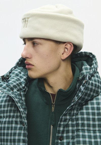 Jeune homme en veste à carreaux verts et pull zippé vert foncé, portant un chapeau blanc doux, regardant vers la gauche sur un fond uni.
