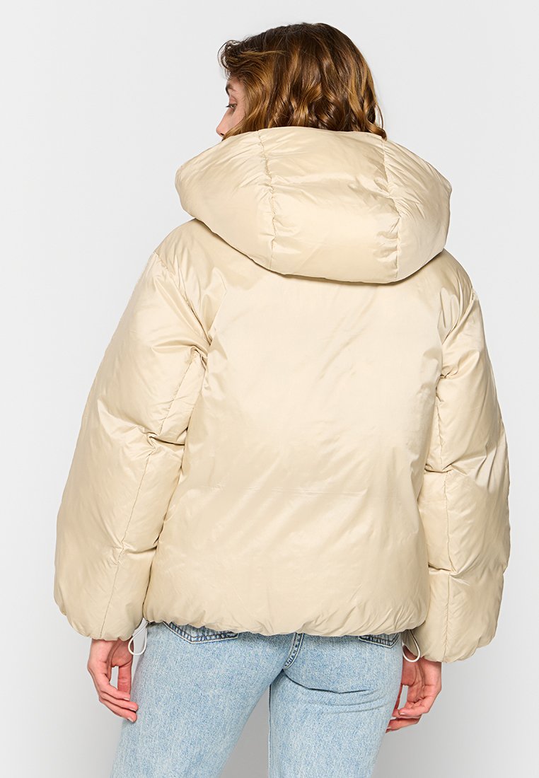 Chaqueta acolchada beige con una gran capucha, mangas oversized y cintura ajustada. Tejido suave y brillante con costuras y detalles de puntadas mínimos.