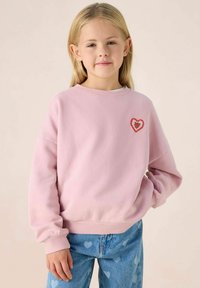Sweatshirt rose clair avec un col rond et des poignets côtelés, orné d'un emblème de cœur rouge sur le côté gauche. Associé à un jean bleu avec des motifs en forme de cœur.