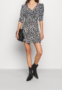 Černobíle vzorované šaty s výstřihem do V, tříčtvrtečním rukávem, přiléhavým pasem a rozšířenou sukní, nošené s černými koženými kotníkovými botami na podpatku a kabelkou.