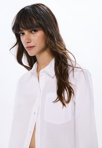Camicia bianca con bottoni realizzata in tessuto leggero, dotata di tasca frontale, colletto classico e vestibilità leggermente ampia. Texture sottile visibile.