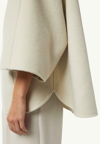 Garment poncho in pile bianco con maniche ampie, caratterizzato da un orlo curvo e una texture liscia. Indossato sopra pantaloni chiari.
