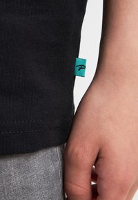 Zwarte geribbelde t-shirt met een turquoise merklabel. Close-up van de zoom en een hand die tegen een lichtgrijze stof rust.
