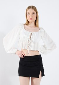 Valge lühikeste varrukatega pluus puffvarrukatega ja paela detailiga, kombineeritud musta mini-seelikuga, millel on külgmine lõhik. Sile, kerge kangas.