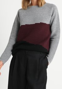 Pull tricoté avec une texture côtelée, partie supérieure grise, milieu bordeaux et sections inférieures noires. Col rond et manches longues.