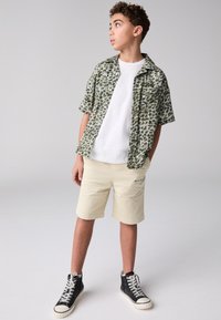 Kortärmad skjorta med knäppning och grönt lövmönster, vit t-shirt, beige shorts och svarta höga sneakers med vita sulor.