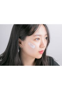 Närbild av ansikte med kräm applicerad på kinden. Långt svart hår, hängande örhängen med kors, naturlig sminkning, mjuk hudstruktur, neutral bakgrund.