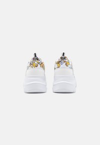 Λευκά αθλητικά παπούτσια με υφή στο πάνω μέρος που παρουσιάζει ένα λουλουδάτο σχέδιο σε κίτρινες και γκρί αποχρώσεις, και μια έντονη μαύρη λαβή στο πίσω μέρος.