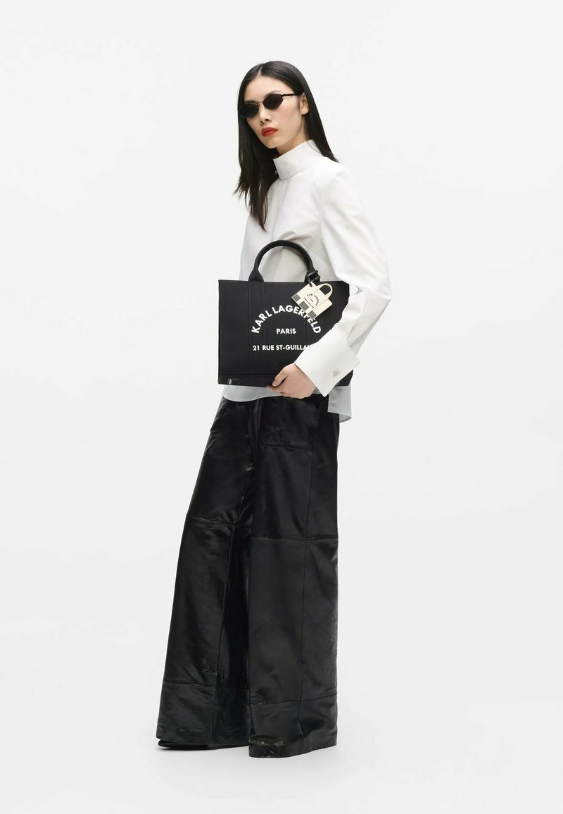 Femme portant des lunettes de soleil noires, un haut blanc à col montant, un pantalon large noir, tenant un sac cabas noir Karl Lagerfeld avec un texte blanc.