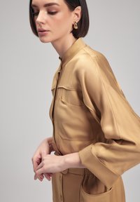 Vestido camisero de satén color marrón claro con un alto cuello, dos bolsillos en el pecho y cierre de botones. Mangas largas con puños enrollados.