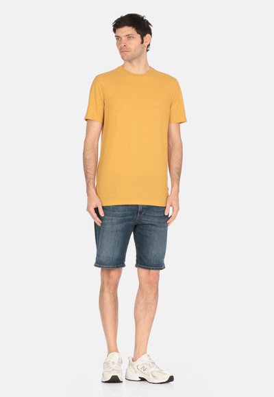 Camiseta de algodón amarilla mostaza con mangas cortas, combinada con pantalones cortos de mezclilla azul oscuro y zapatillas deportivas de color claro. Diseño sencillo, sin patrones.