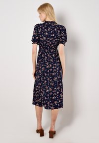 Marineblaues Blumenkleid in Midi-Länge mit kurzen Puffärmel, gerafftem Bund und einem schwingenden Rock, der mit rosa und weißen Blumenmustern verziert ist. Quadrat-Ausschnitt.