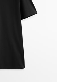T-shirt noir à manches courtes suspendu sur un fond blanc immaculé, montrant la manche droite et une partie du torse.