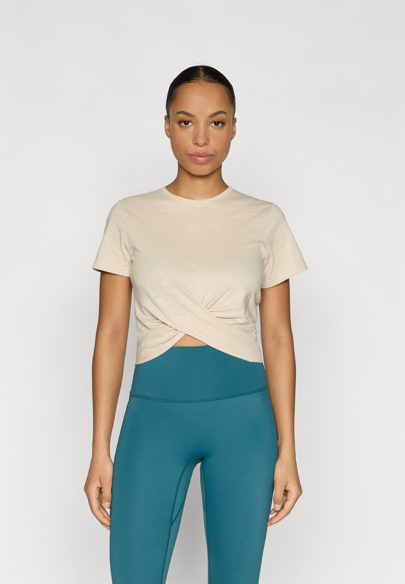 Μπεζ κοντό t-shirt με σχέδιο μπροστά με στρίψιμο, κοντά μανίκια, στρογγυλή λαιμόκοψη, συνδυασμένο με ψηλόμεσα τιρκουάζ κολάν. Λείο υλικό.