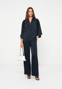 Marineblaue Seidenbluse mit weiten Ärmeln und einer Knopfleiste vorne, kombiniert mit passenden weit geschnittenen Hosen; hält eine kleine weiße Handtasche.