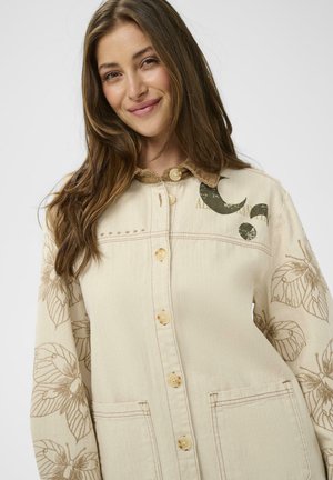 Ung kvinde med langt brunt hår iført en beige jakke med knapper, dekoreret med blomsterprint på ærmerne og en manchet i kordfløjl, som smiler let.