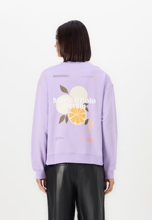 Kvinde med skulderlangt mørkt hår iført en lavendelfarvet sweatshirt med grafik af citrusfrugter og teksten "Marc O'Polo Denim" på ryggen.