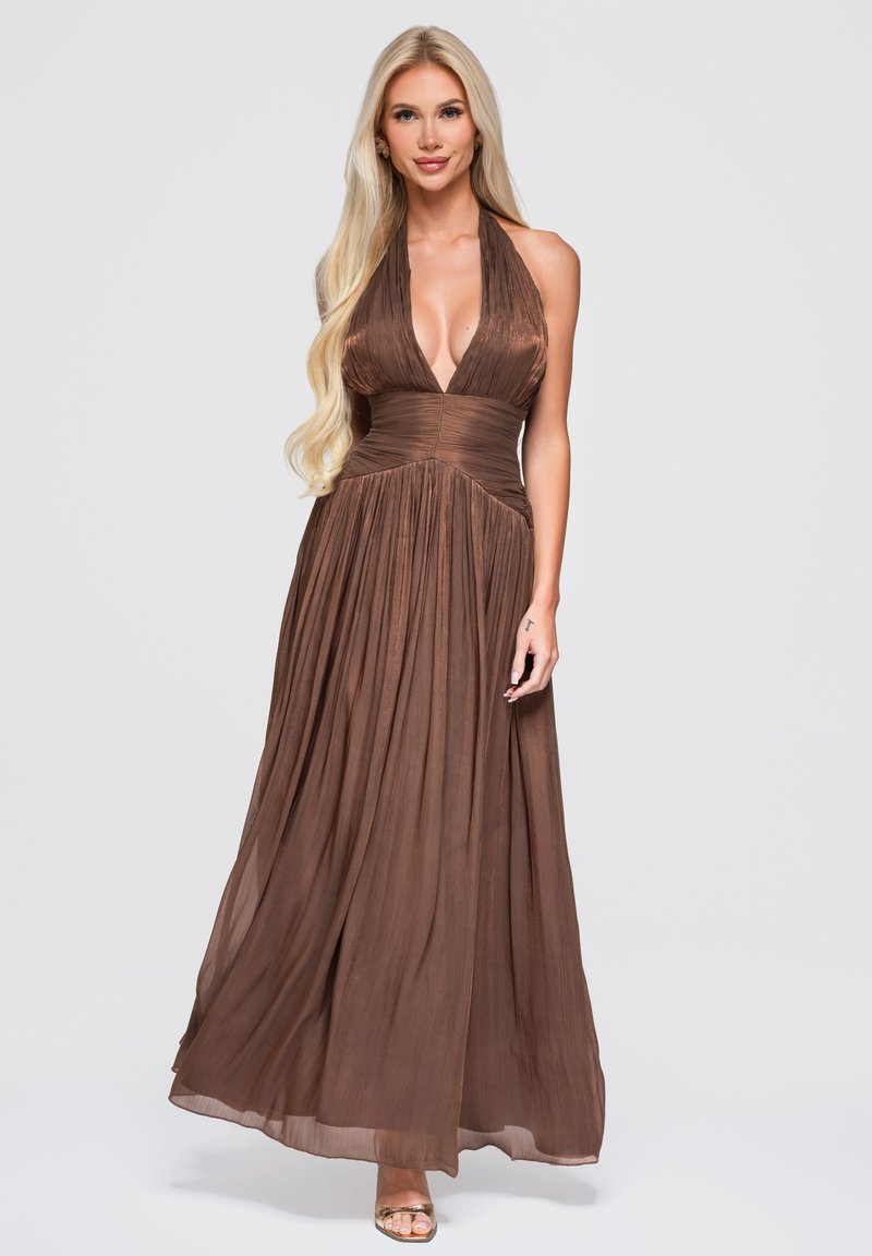 Femme blonde portant une longue robe marron fluide sans manches avec un profond décolleté en V, debout devant un fond uni gris clair.