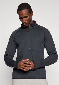 Pull-over noir à demi-zip en matériau texturé avec un motif subtil. Doté d'un poignet élastique et d'une poche zippée sur le côté.