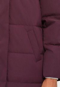 Detailný záber ruky pri vrecku fialovej zimnej bundy s vycpávkou a vodorovným prešívaním.
