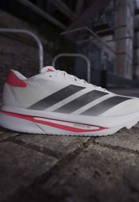 Hvid og pink Adidas løbesko med sorte striber på brostensbelægning i et urbant miljø.