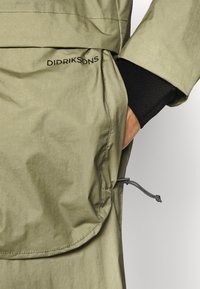 Oliwkowa kurtka o gładkiej, wodoodpornej fakturze. Posiada dużą boczną kieszeń, ściągacz na sznurku oraz czarne siateczkowe wykończenie mankietów.