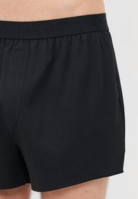Black men's boxer shorts with elastic waistband and subtle brand logo worn on lower torso and upper thighs.