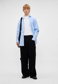 Világoskék csíkos, gombos ing fehér póló fölött, fekete kordnadrággal párosítva. Kiegészítők között láncos nyaklánc és postás táska van.