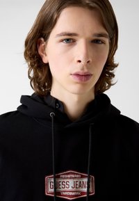 Czarna bluza z kapturem z szarymi sznurkami i teksturowanym logo "Guess Jeans" w kolorach czerwono-białych. Wykonana z miękkiego materiału o luźnym kroju.