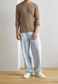 Brun stickad tröja med ribbat mönster, rund hals och uppvikta ärmar, parat med ljusblåa löst sittande jeans och vita sneakers på trägolv.