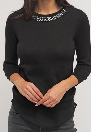 Kvinde iført en sort langærmet sweater med perlebesat halsudskæring, hænderne foldet ved taljen, kombineret med mørkegrå bukser.