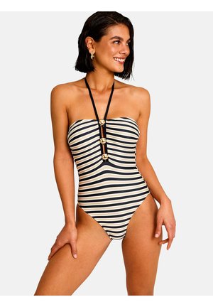 Jeune femme portant un maillot de bain sans bretelles à rayures horizontales noires et blanches avec trois boutons dorés et une bride halter noire, souriante.