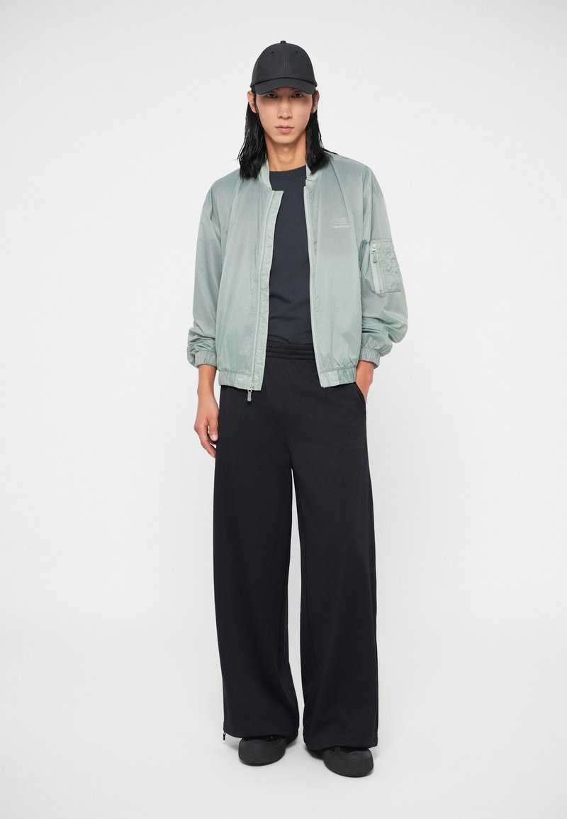 Giacca bomber verde chiaro con zip, con polsini elasticizzati, abbinata a ampi pantaloni neri e un cappellino nero. Tessuto morbido, design semplice.