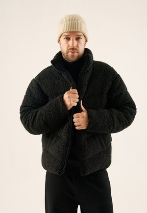 Schwarze Fleecejacke aus strukturiertem Stoff, hoher Kragen und Reißverschluss. Kombiniert mit einem schwarzen Rollkragenpullover und einer beigen Strickmütze.