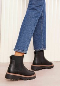 Botas de tobillo de cuero negro con paneles laterales elásticos, suela texturizada y una suela marrón gruesa, combinadas con unos jeans de denim azul.