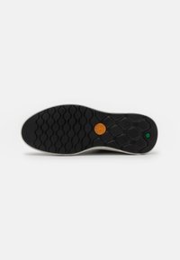 Semelle de chaussure en caoutchouc noire avec un motif hexagonal, comportant un logo circulaire orange et un point d'accent vert. Bords lisses et arrondis visibles.