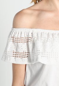 White off-shoulder top featuring a ruffled lace overlay with cut-out patterns and elastic neckline. Soft fabric, plain body design.