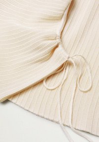 Top en maille côtelée couleur beige clair avec un détail froncé à l'ourlet, comportant un cordon de serrage et une texture douce pour un ajustement réglable.