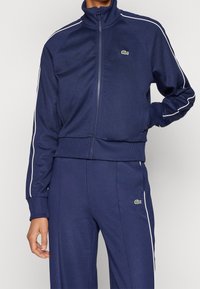 Chándal azul marino con franjas blancas, con cuello alto y puños acanalados. Logo de Lacoste en el pecho izquierdo y en los pantalones.