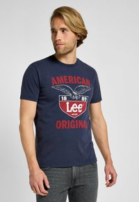 Camiseta de algodón de color azul marino con un gran diseño gráfico que presenta letras en rojo y blanco y un emblema de águila. Mangas cortas y cuello redondo.