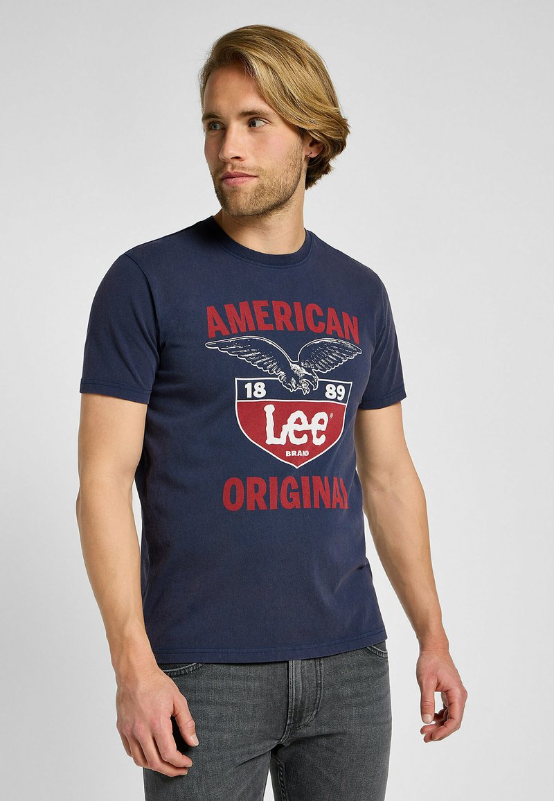 Camiseta de algodón de color azul marino con un gran diseño gráfico que presenta letras en rojo y blanco y un emblema de águila. Mangas cortas y cuello redondo.