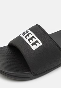 Fekete csúszásmentes szandál, texturált pánttal, amelyen fehér "REEF" logó található. A talpa enyhén mintázott a tapadás és a kényelem érdekében.
