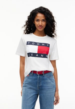 Kvinde med krøllet hår iført en hvid Tommy Jeans t-shirt, blå jeans og et rødt bælte, stående mod en hvid baggrund.