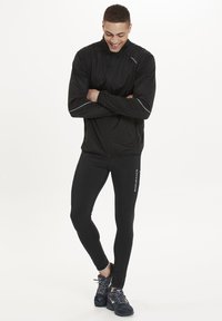 Schwarze leichte Jacke mit reflektierenden Akzenten, kombiniert mit schwarzen Leggings mit dem Schriftzug "ENDURANCE". Das Model trägt dunkle Sportschuhe.