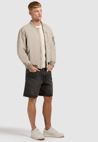 Homme portant une veste bomber beige, un t-shirt blanc, un short en denim noir et des baskets blanches, debout les mains dans les poches devant un fond uni.