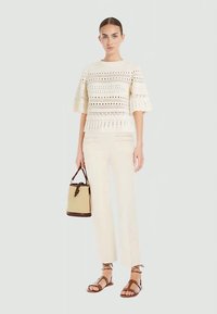 Moteris vilki grietinės spalvos nėriniuotą viršų, grietinės spalvos kelnes, rudus šniūrinius sandalus ir laiko kreminės bei rudos spalvos rankinę.