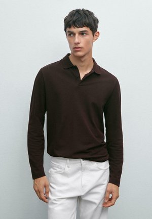 Giovane uomo che indossa una polo a maniche lunghe marrone scuro e pantaloni bianchi, guarda di lato su uno sfondo chiaro e uniforme.